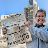 Ein Pfarrer steht vor einer eingerüsteten Kirche und hält eine alte Zeitung in den Händen.