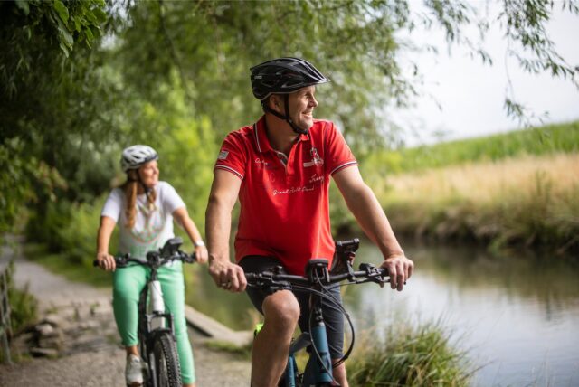 Ein Mann und eine Frau fahren mit dem Fahrrad an einem Fluss vorbei.