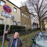 Ein Mann mit Schal zeigt auf ein Halteverbotsschild in einer ruhigen Wohnstraße.