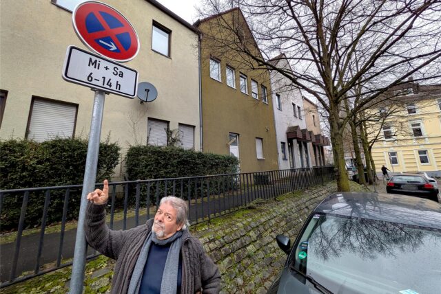Ein Mann mit Schal zeigt auf ein Halteverbotsschild in einer ruhigen Wohnstraße.