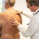 Ein oberkörperfreier Mann mittleren Alters steht mit dem Rücken zum Arzt, der in weißem Kittel den Rücken untersucht.
