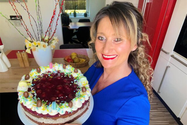 Eine lächelnde Frau präsentiert stolz eine selbstgebackene Kirschtorte, die mit Schlagsahne, Schokoeiern und bunten Streuseln verziert ist. Im Hintergrund sind frühlingshafte Dekorationen wie blühende Zweige und Narzissen zu sehen, die auf eine festliche Atmosphäre hinweisen.