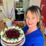 Eine lächelnde Frau präsentiert stolz eine selbstgebackene Kirschtorte, die mit Schlagsahne, Schokoeiern und bunten Streuseln verziert ist. Im Hintergrund sind frühlingshafte Dekorationen wie blühende Zweige und Narzissen zu sehen, die auf eine festliche Atmosphäre hinweisen.