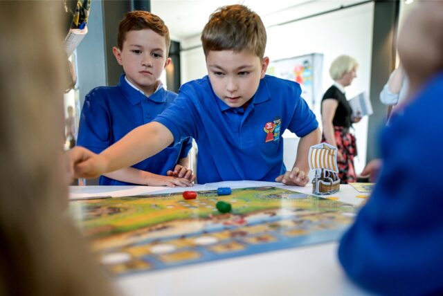 Zwei Jungen in blauen Poloshirts spielen ein Brettspiel. Das Spiel enthält eine Spielfigur in Form eines Wikingerschiffs und farbige Spielsteine.