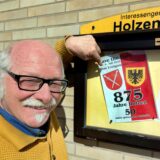 Ein Mann zeigt auf eine Einladung zu einer Jubiläumsfeier in einem Schaukasten an einer Hauswand.
