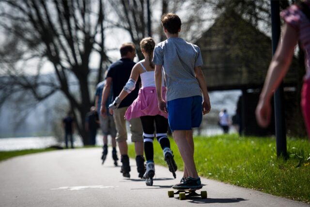 Eine Gruppe von Personen, darunter ein Junge auf einem Skateboard, fährt auf einem Bürgersteig entlang, einige von ihnen mit Inlineskates. Die Gruppe ist von hinten zu sehen, während sie sich durch eine parkähnliche Umgebung bewegt.