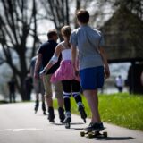 Eine Gruppe von Personen, darunter ein Junge auf einem Skateboard, fährt auf einem Bürgersteig entlang, einige von ihnen mit Inlineskates. Die Gruppe ist von hinten zu sehen, während sie sich durch eine parkähnliche Umgebung bewegt.