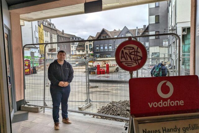 Ein Einzelhändler steht vor einem Bauzaun, der den Zugang zu seinem Geschäft verbarrikadiert.
