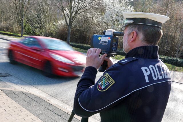 Ein Polizist kontrolliert die Geschwindigkeit, ein rotes Auto fährt an ihm vorbei.