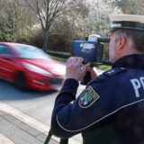 Ein Polizist kontrolliert die Geschwindigkeit, ein rotes Auto fährt an ihm vorbei.