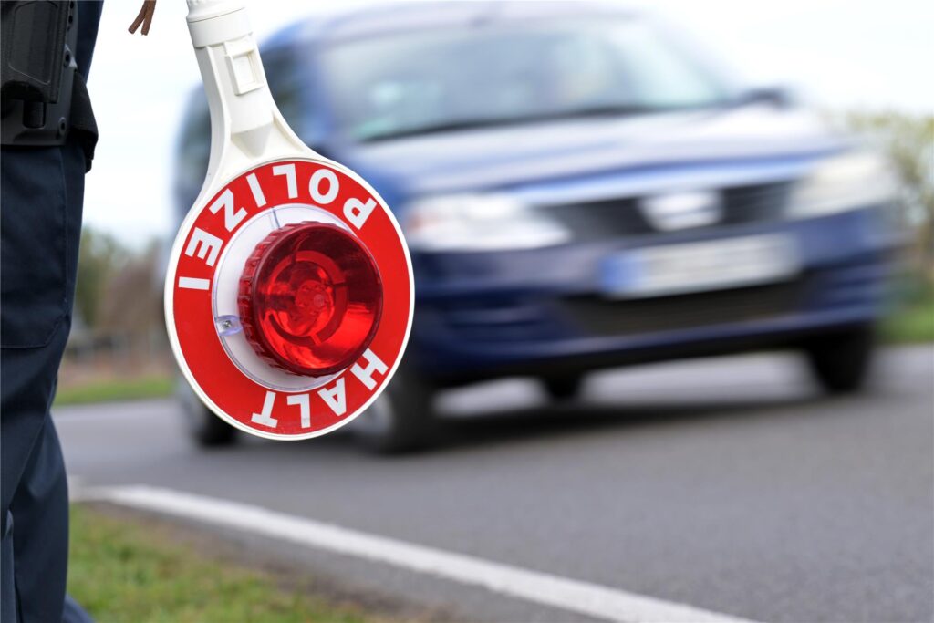 Eine Person steht mit einer Verkehrskelle am Straßenrand, ein blaues Auto fährt über die Straße.