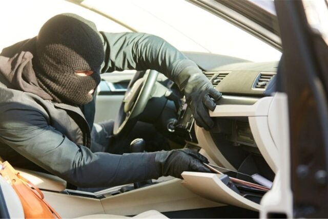 Eine dunkle Gestalt mit Sturmhaube und Lederhandschuhen durchsucht das Handschuhfach eines Autos.