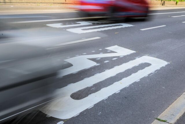Auf dem Bild ist ein Autofahrzeug zu sehen, das sich auf einer Straße mit einer Geschwindigkeitsbegrenzung von 30 km/h bewegt. Die weiße Markierung auf der Fahrbahn signalisiert die Geschwindigkeitsbegrenzung.