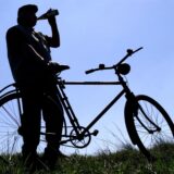 Ein Mann trinkt aus einer Flasche, während er sich auf ein Fahrrad auf einer Wiese lehnt. Die Silhouette des Mannes und des Fahrrads heben sich vom hellblauen Himmel ab.