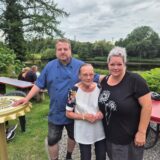 Drei Personen posieren für ein Foto im Freien, mit einem Fluss und Bäumen im Hintergrund. Im Vordergrund sind Tische und Stühle zu sehen, was auf einen Garten oder eine Terrasse hindeutet.