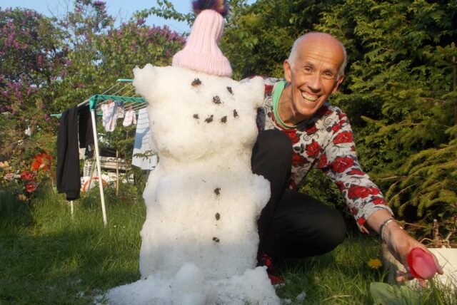 Ein 65-jähriger schlanker Mann sieht bei Sonnenschein und offenbar warmen Temperaturen neben einem Schneemann.