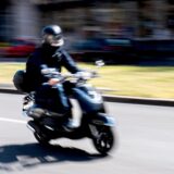 Eine Person mit Helm fährt mit einem Motorroller auf einer Straße.