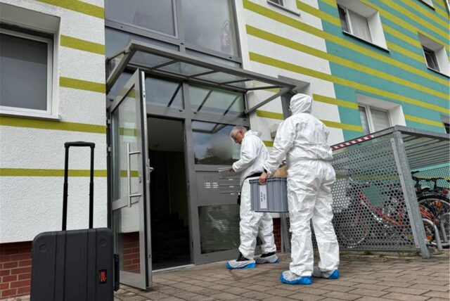 Zwei Menschen in weißen Schutzanzügen stehen vor einem Mehrfamilienhaus. Einsatzkräfte untersuchen den Tatort, an dem eine 43-Jährige erstochen wurde.