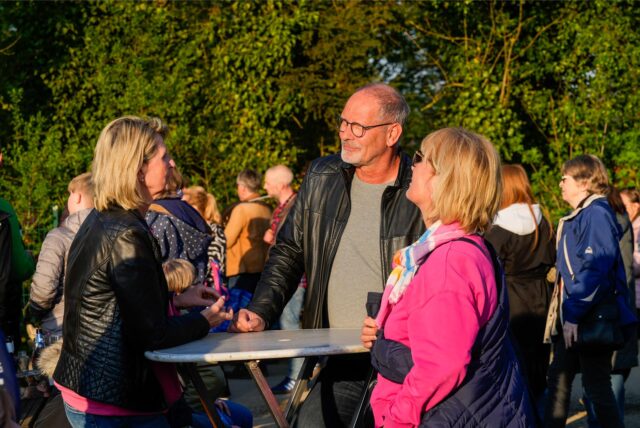Zwei Frauen und ein Mann stehen rund um einen Stehtisch. Im Hintergrund sind weitere Menschen zu sehen.