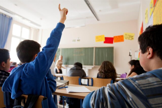 Schulkinder sitzen in einem Klassenraum. Ein Junge zeigt auf.