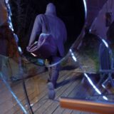 Im Vordergrund eine zerschlagene Terrassentür, im Hintergrund eine dunkel gekleidete Person mit einer Sporttasche über der Schulter.