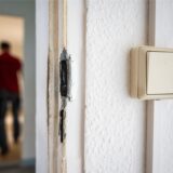 Das zerstörte Schloss einer Wohnungstür, im Hintergrund unscharf eine Person.