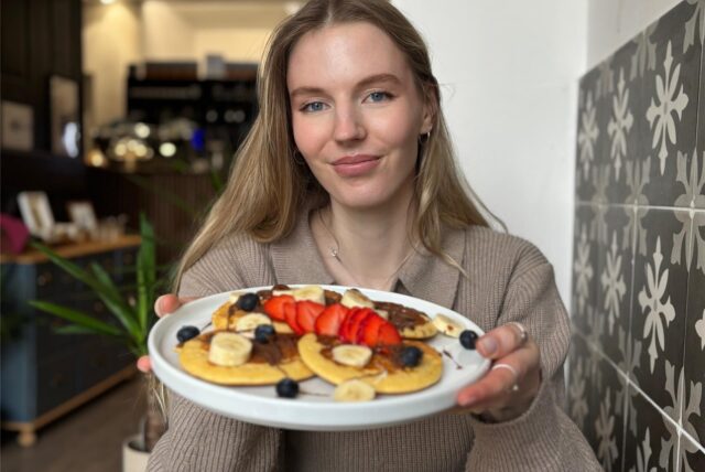 Eine junge Frau präsentiert einen Teller mit Pancakes, die mit Schokoladenaufstrich, Bananen, Erdbeeren und Blaubeeren belegt sind. Die Szene spielt sich vermutlich in einem Café ab, wie die Einrichtung im Hintergrund vermuten lässt.