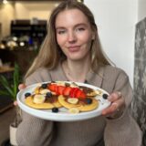 Eine junge Frau präsentiert einen Teller mit Pancakes, die mit Schokoladenaufstrich, Bananen, Erdbeeren und Blaubeeren belegt sind. Die Szene spielt sich vermutlich in einem Café ab, wie die Einrichtung im Hintergrund vermuten lässt.