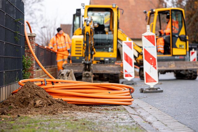 Arbeiter sind mit Bagger und Spaten an einer Straße mit der Erdverlegung von Glasfaserkabeln beschäftigt.