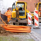 Arbeiter sind mit Bagger und Spaten an einer Straße mit der Erdverlegung von Glasfaserkabeln beschäftigt.