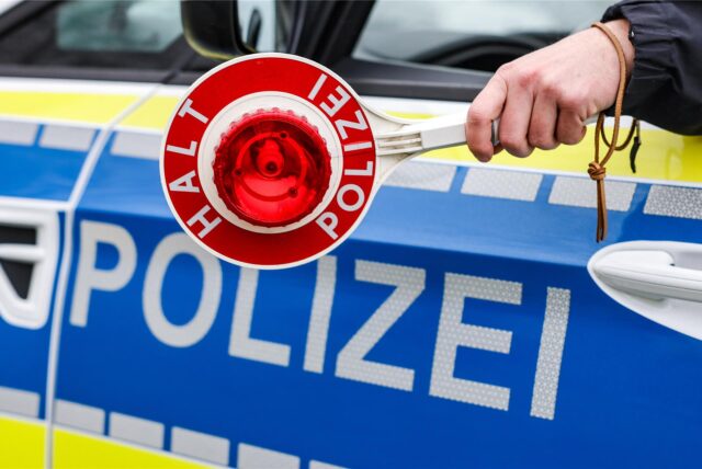 Eine Hand hält eine Polizeikelle, im Hintergrund ein Streifenwagen.