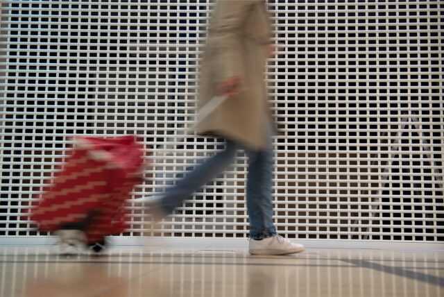 Eine Person in einer beigefarbenen Jacke zieht einen roten Einkaufstrolley hinter sich her.