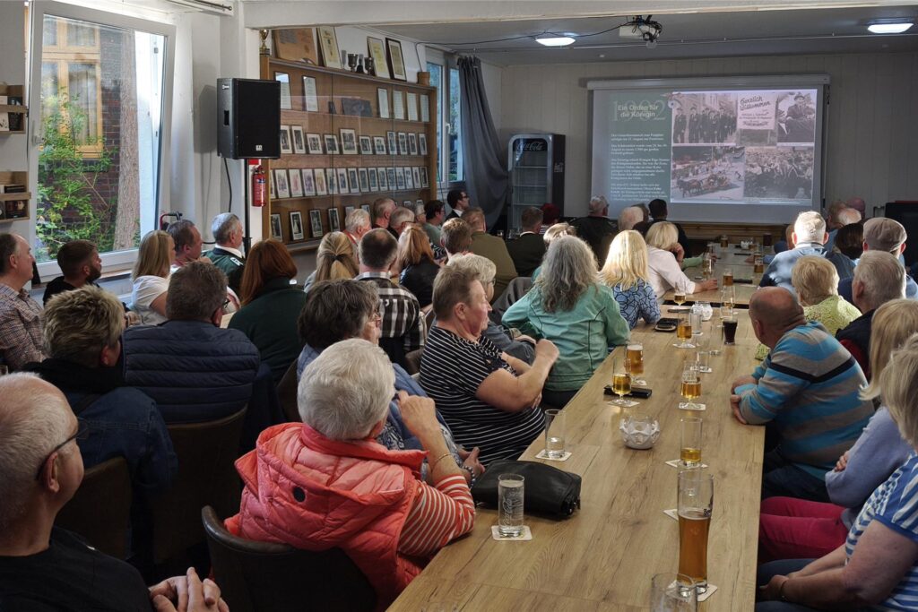 Eine Gruppe von Menschen sitzt an einem langen Tisch und schaut auf eine Leinwand, auf der alte Fotografien der Bürgerschützengilde St. Stephanus Bork und Text projiziert werden. Getränke stehen auf dem Tisch und ein Regal mit Fotos ist an der Wand im Hintergrund zu sehen.