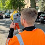 Vermessungsingenieur Peter Pohl und ein Kollege stehen in Signaljacken mit Nivelliergerät und Klemmbrett an der Friedrich-Ebert-Straße in Unna, um in etwa 15 Metern Entfernung eine Messlatte anzuvisieren.