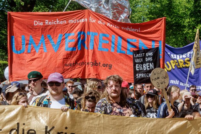 Teilnehmer halten auf einer Mai-Demo mit dem Motto „Superreiche? Superscheiße!“ ein Transparent mit der Aufschrift „Umverteilen!!“.