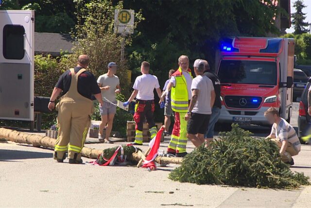 Der umgestürzte Maibaum liegt auf dem Boden. Krankenwagen steht im Hintergrund und Einsatzpersonal läuft herum.