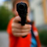 Eine vorgehaltene Pistole im Vordergrund, ganz unscharf, dahinter ein Jugendlicher.