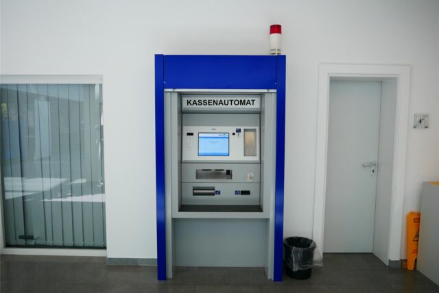 Ein Kassenautomat in einem Innenraum wird gezeigt. Der Automat ist blau umrandet und befindet sich zwischen einer Tür und einer Fensterfront.