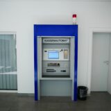 Ein Kassenautomat in einem Innenraum wird gezeigt. Der Automat ist blau umrandet und befindet sich zwischen einer Tür und einer Fensterfront.