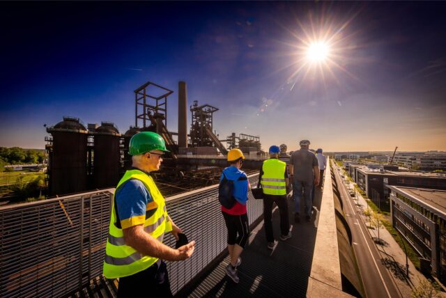 Eine Besuchergruppe geht im Sonnenlicht über den Skywalk. Im Hintergrund ist die denkmalgeschützte Hochofenanlage zu sehen.