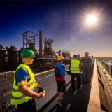 Eine Besuchergruppe geht im Sonnenlicht über den Skywalk. Im Hintergrund ist die denkmalgeschützte Hochofenanlage zu sehen.