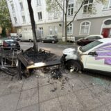 Verkohlter Sperrmüll liegt auf dem Gehweg. Ein nebenstehendes Auto ist von dem Brand an der Front erheblich beschädigt worden. Ein Fenster ist mit Folie abgeklebt.