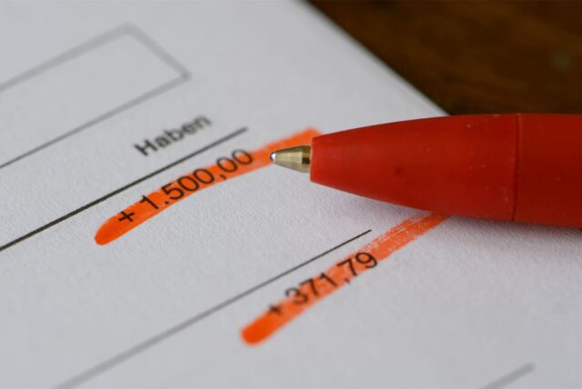 Der vordere Teil eines orangefarbenen Kugelschreibers liegt auf einem Blatt Papier. Darauf steht „Haben“, darunter folgt ein Strich. Darunter ist „+1.500,00“ zu lesen. Nach einer Lücke folgt ein weiterer Strich und darunter „+371,79“. Die beiden Zahlen sind rot markiert.
