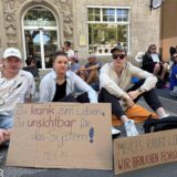 Etwa 100 Betroffene und Angehörige der Multisystemerkrankung ME/CFS demonstrienden im Liegen vor der Reinoldikirche am Ostenhellweg in Dortmund, Eine Frau hält das Plakat hoch mit der Aufschrift: „Wir fordern Inklusion und Forschung ME/CFS“.
