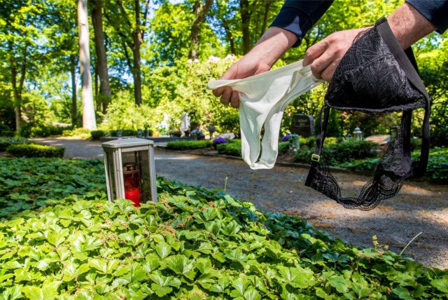 Auf dem Waldfriedhof in Herten sind über einem Grab zwei Hände zu sehen, die einen weißen Damenslip und einen schwarzen BH halten.