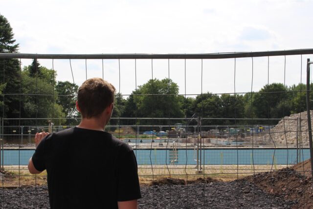 Ein junger Mann beobachtet die Baustelle zum neuen Sesekebad in Kamen durch einen Bauzaun hindurch.