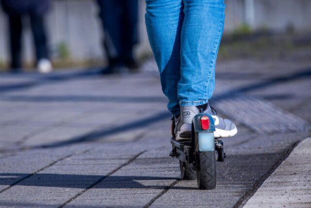Eine Person fährt mit einem E-Scooter. Nur ein Teil der Hose und die Schuhe auf dem Fahrzeug sind zu sehen.