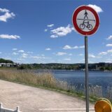 Ein Schild, das Fußgängern und Radfahrern den Weg verbietet, steht an einem Weg entlang eines Sees unter einem blauem Himmel mit Wolken. Im Hintergrund sind Häuser und ein bewaldeter Hügel zu sehen, während Spaziergänger den Weg nutzen.
