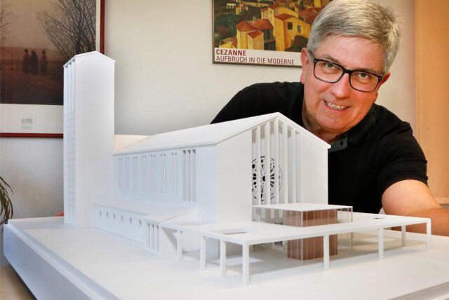 Hans-Jürgen Klein aus dem Kirchenvorstand der katholischen Gemeinde Bergkamen schaut hinter dem Model einer Kirche hervor.
