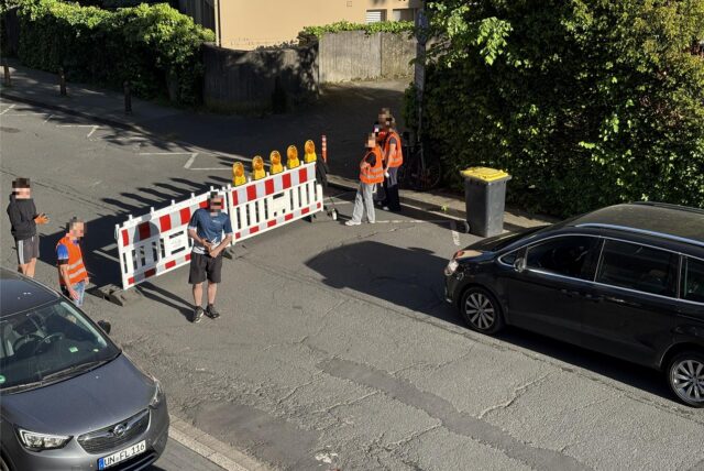 Ein schwarzes Auto steht vor einer Absperrung, an der mehrere Ordner teils in orangefarbenen Warnwesten stehen